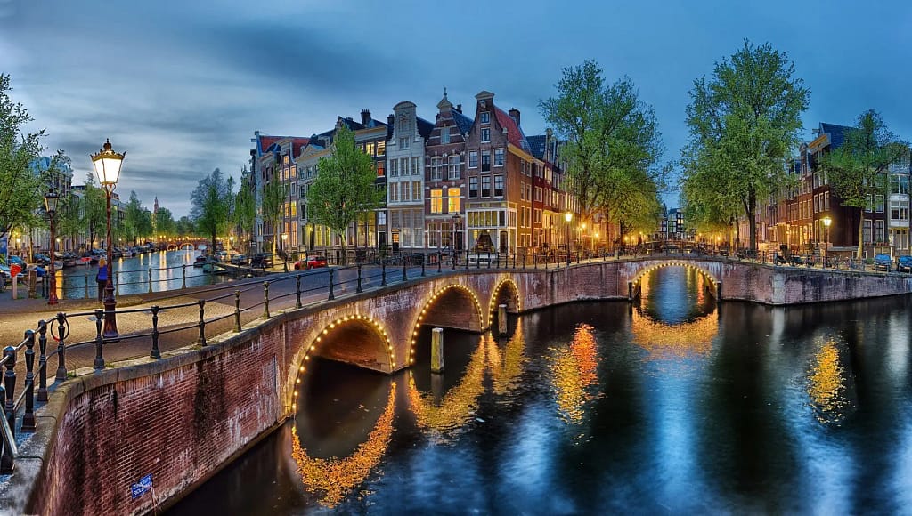 Scenic view of Amsterdam canal with illuminated bridges and traditional Dutch buildings at dusk.