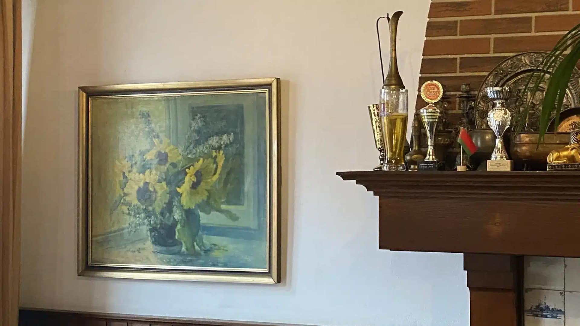 Framed painting of sunflowers on a wall with a mantelpiece displaying trophies and decorative items in the lounge area at Budget Hotel Sphinx Amsterdam.
