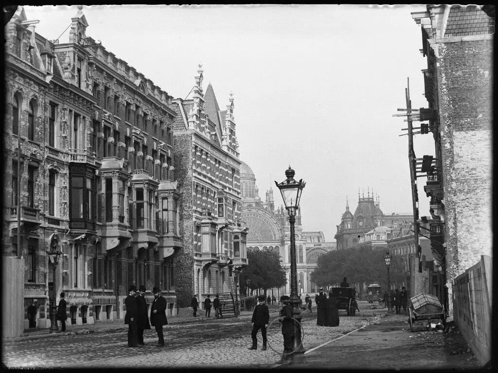 Historic black and white photo of a street in Amsterdam with ornate buildings and people in period clothing